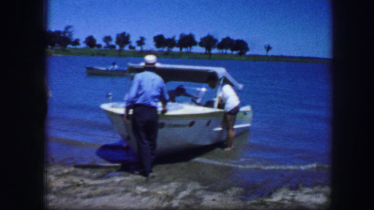 In Fort Worth, Texas, 1964 home-movie footage, two adults stand near a white speedboat in shallow lake water.