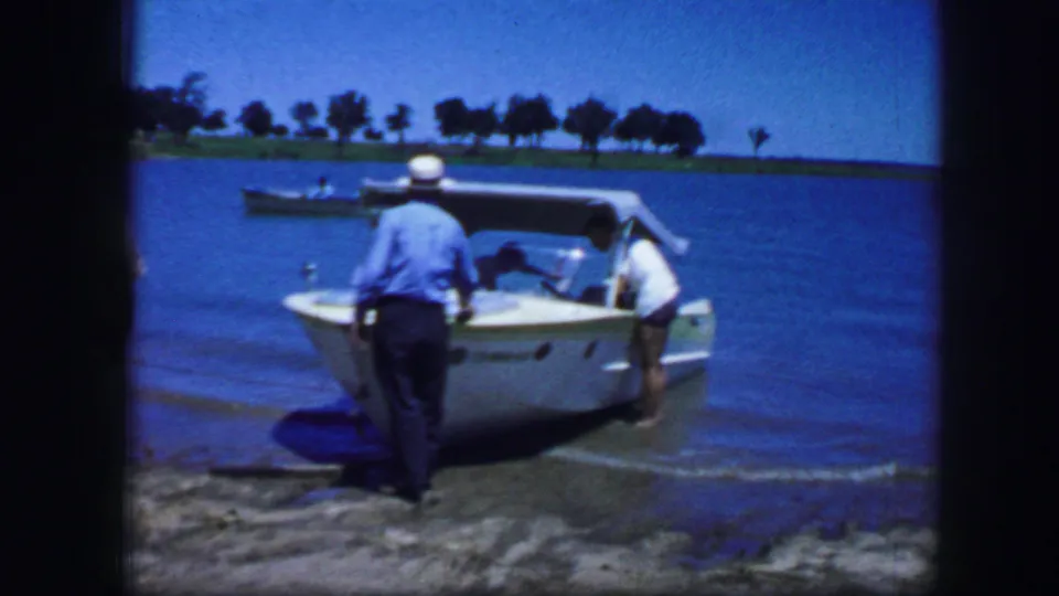 In Fort Worth, Texas, 1964 home-movie footage, two adults stand near a white speedboat in shallow lake water.