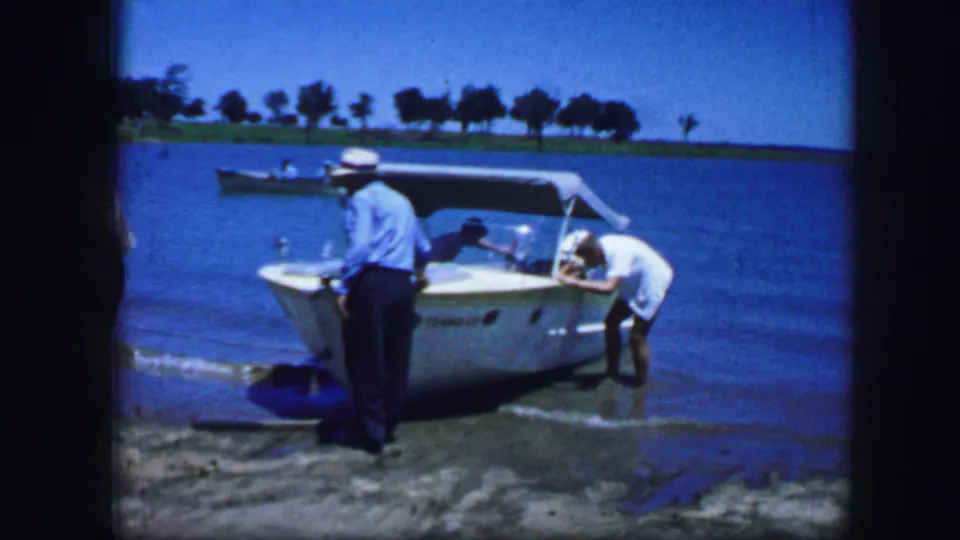A 1964 Fort Worth lakeside frame shows a man at the stern and another adult leaning into a white speedboat near the bow.