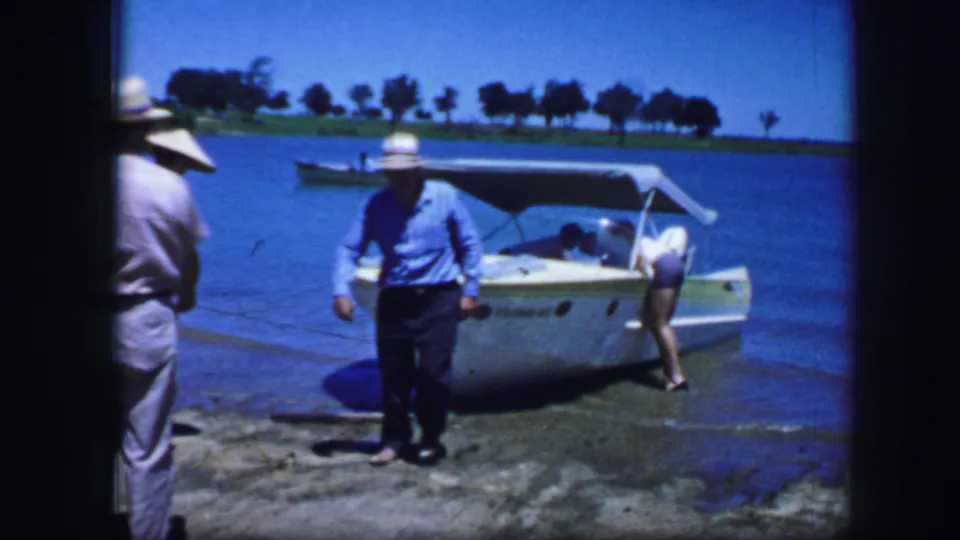 In the 1964 Fort Worth home-movie scene, a man walks from the shoreline while another adult remains beside the speedboat.