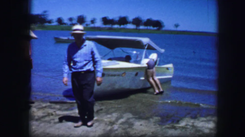 A man in a blue shirt faces the camera as another adult leans over the white speedboat in shallow Fort Worth lake water.