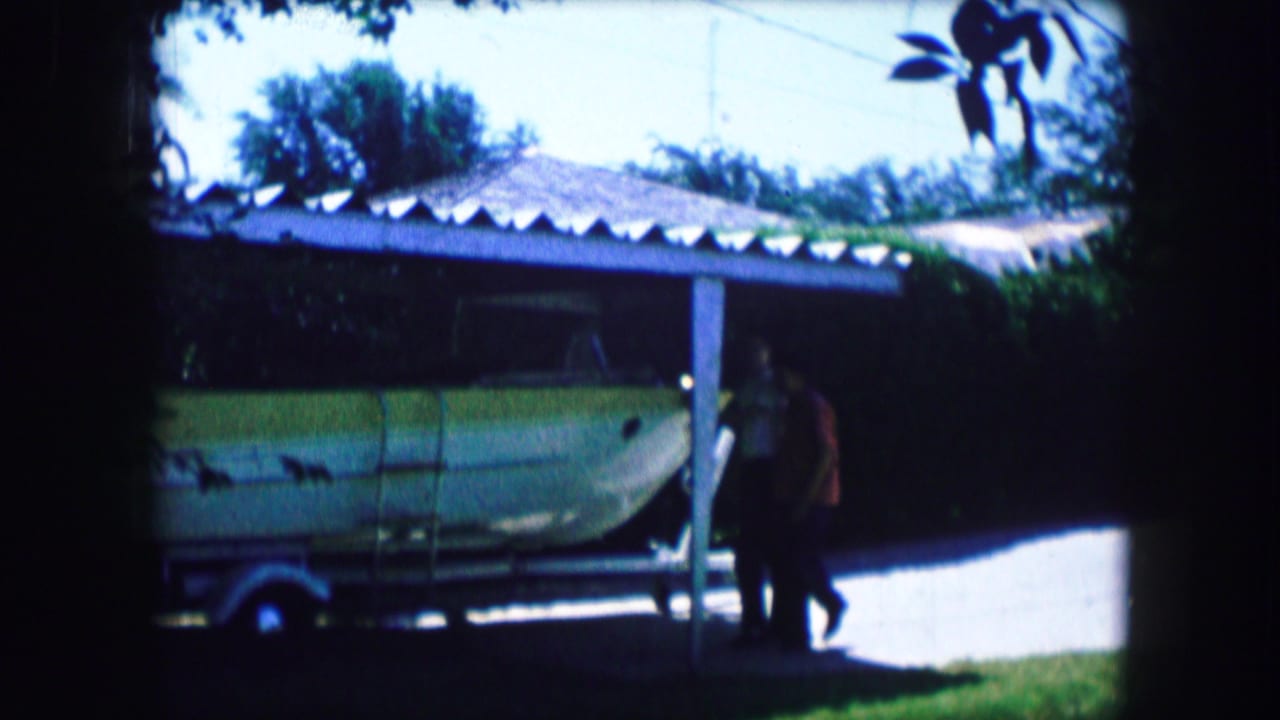 1965 Lewisville, Texas home-movie frame showing two men beside a pale green boat on a trailer outside a house.
