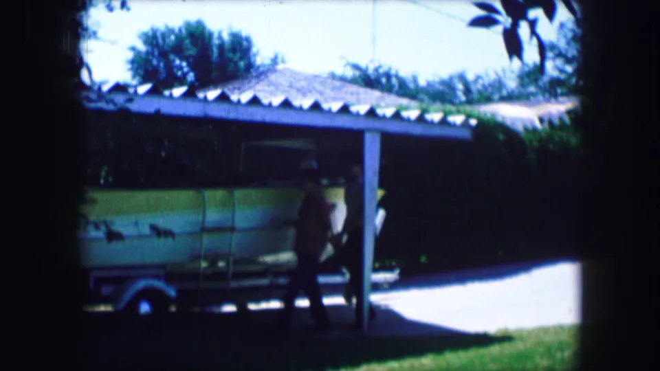A 1965 Texas home-movie view of a man near a pale green boat on a trailer beside a shaded carport.