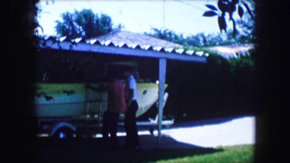 Two adult men appear near the front of a trailered boat outside a Lewisville, Texas house in 1965 home-movie footage.
