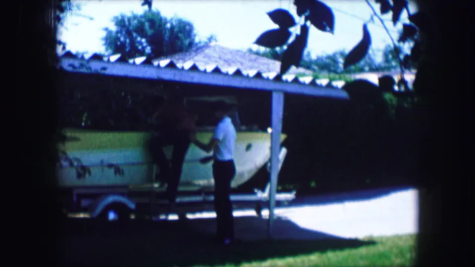 A home-movie frame shows two men beside a pale green boat and trailer in a sunny 1965 Lewisville driveway scene.