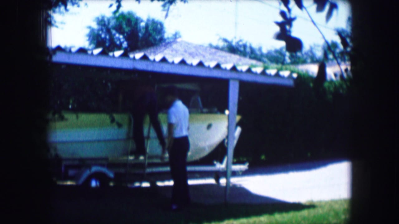 Man standing beside a small aircraft on a trailer under a shaded carport.