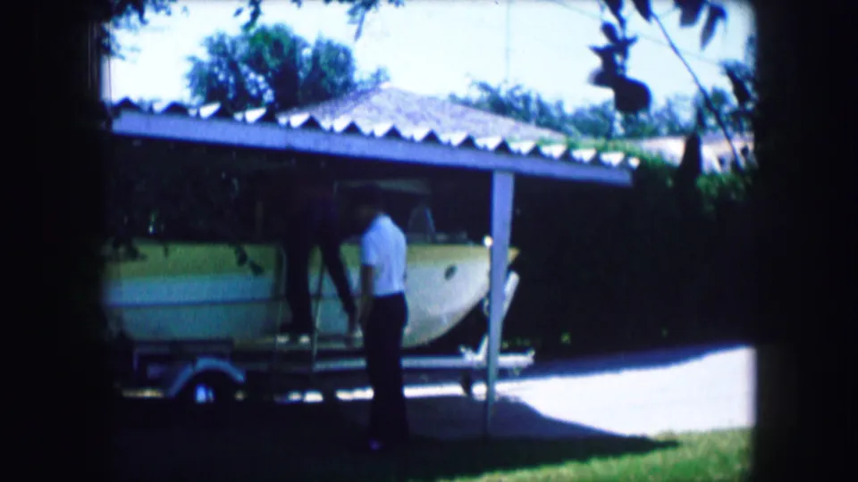 1965 Lewisville, Texas 8mm frame showing a man in a white shirt beside a light-colored small aircraft parked on a trailer near a carport.