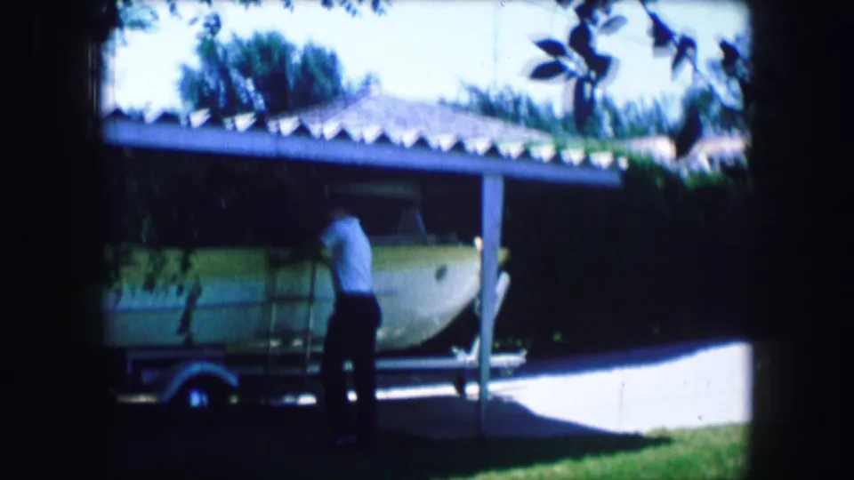 1965 Lewisville, Texas 8mm frame showing the side of a small aircraft and a man leaning or standing close to it near a carport.