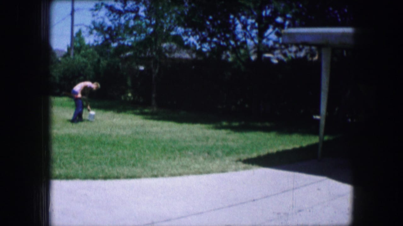 Adult figure bends over on a backyard lawn near a small container or object.