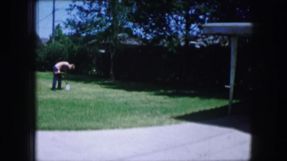 Adult figure remains bent over on a suburban lawn, with driveway, hedges, and trees visible in 1965 Lewisville footage.