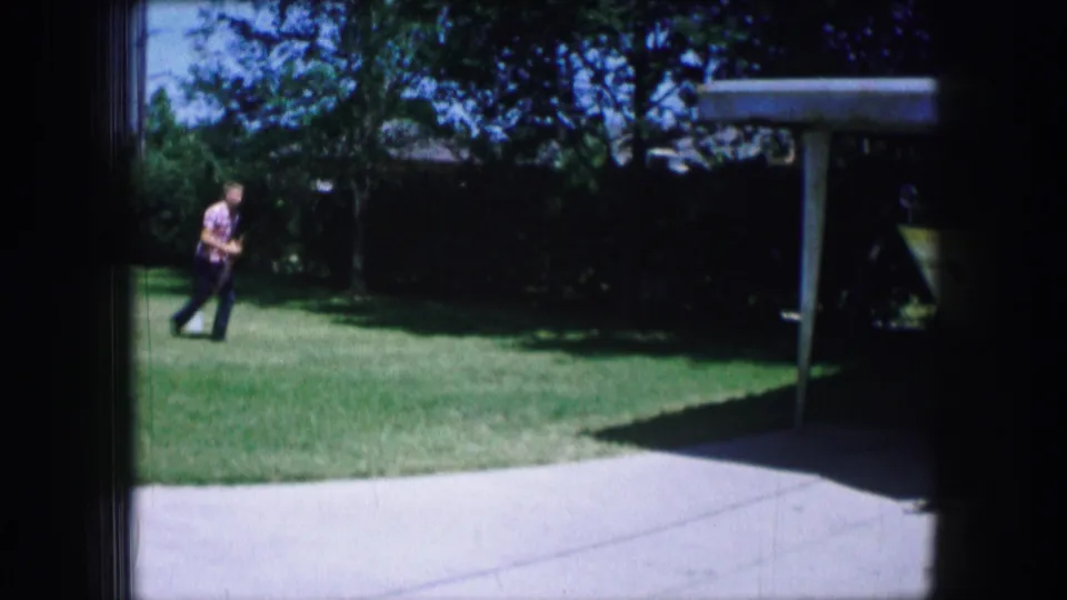Adult man walks across a Lewisville, Texas backyard lawn near the driveway and carport in 1965 8mm footage.