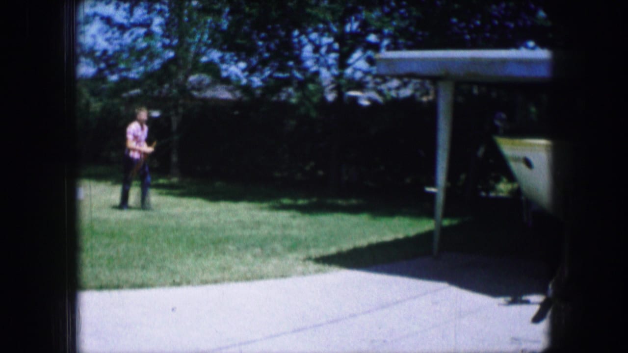 Man walking across a sunlit lawn near a driveway and boat under a carport.