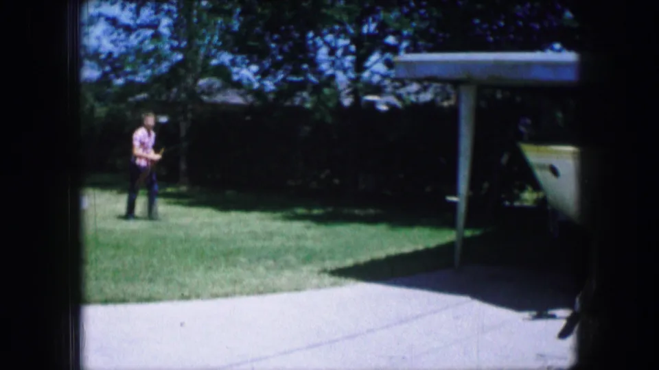 In 1965 Lewisville, Texas, a man in a red shirt walks across a grassy yard beside a driveway and a boat under a carport.