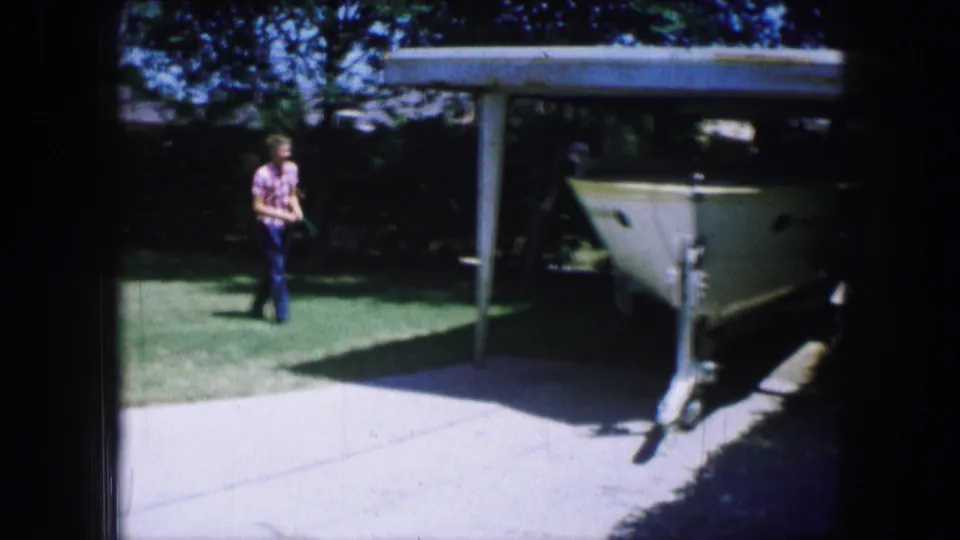 The man is nearer the driveway as the boat, trailer hitch, carport roof, and shaded trees frame the residential yard.