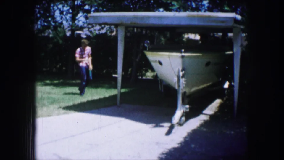 Near the end of the shot, the man is framed beside the lawn and driveway with the boat parked under the carport in bright sun.