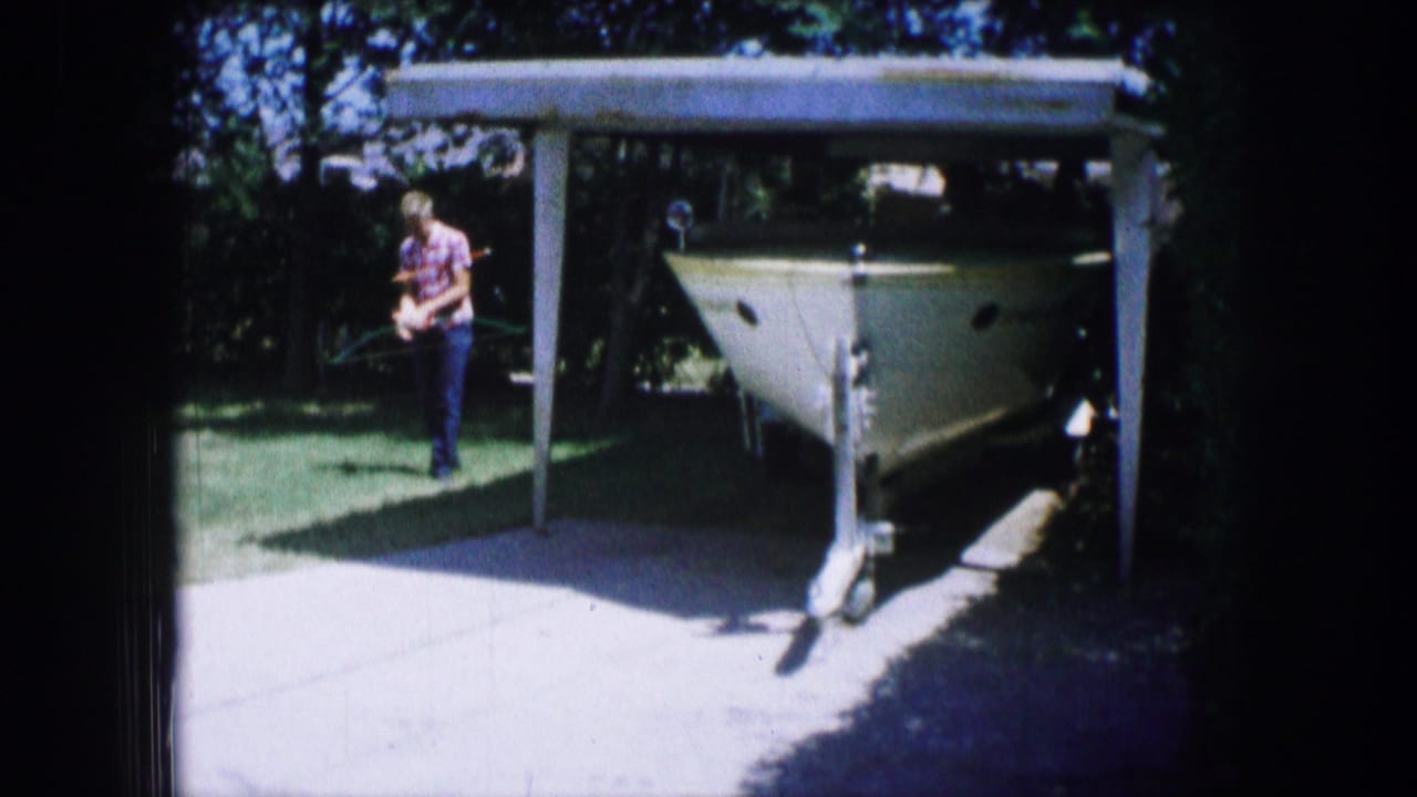 White boat on a trailer under a carport with a person walking nearby on the lawn.
