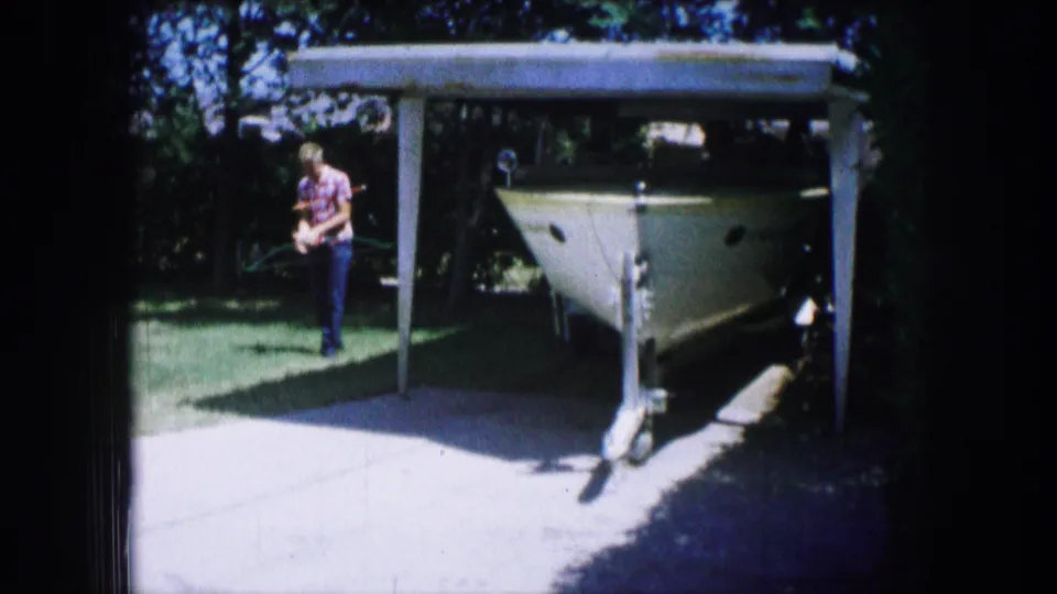1965 Lewisville, Texas 8mm frame showing a white boat under a carport, driveway, lawn, trees, and a person walking at left.