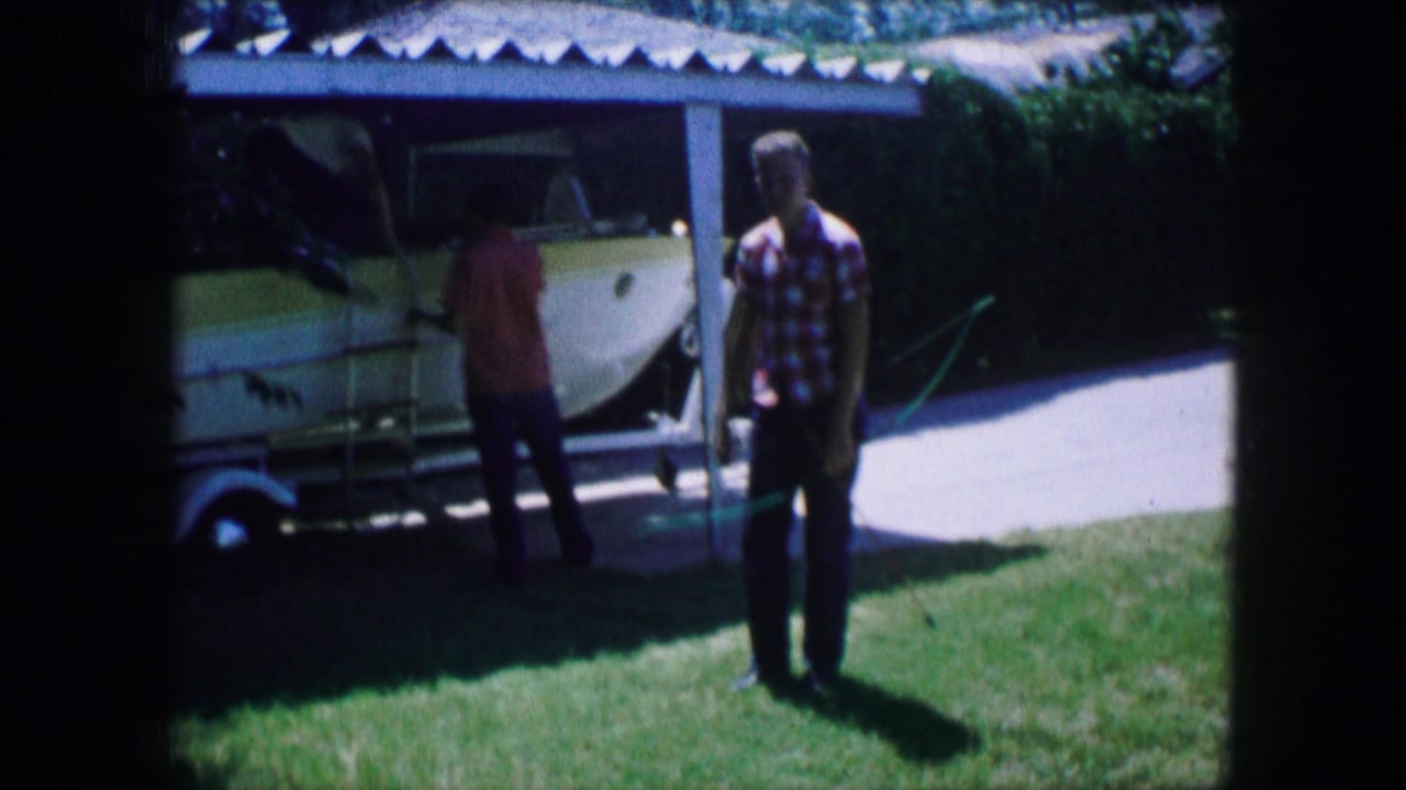Two people stand near a white boat on a trailer beside a driveway and lawn.