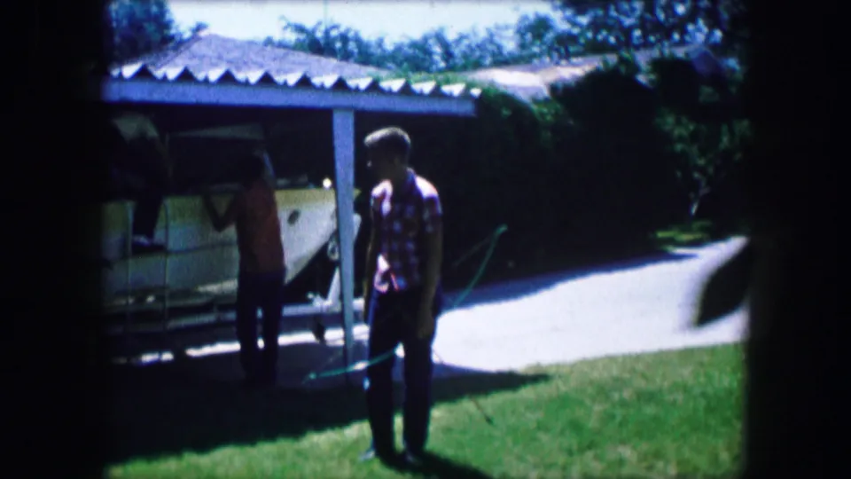 1965 Lewisville, Texas 8mm frame showing one person near the boat and another standing in front of the lawn and driveway.