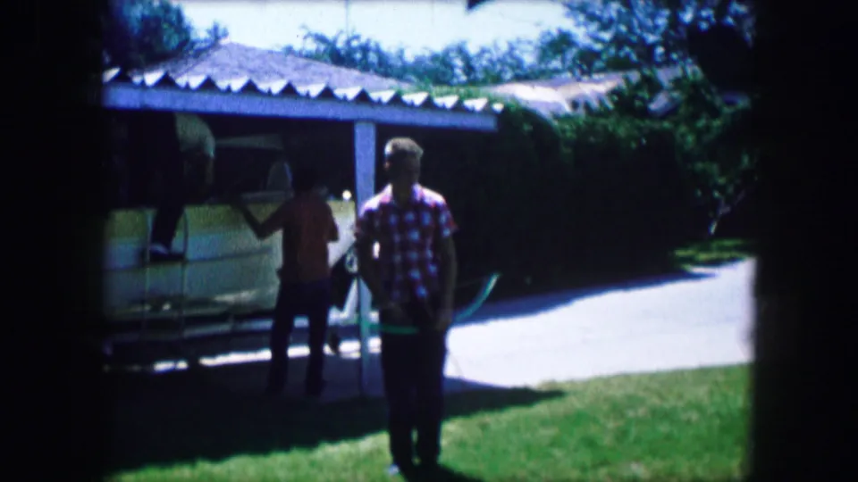 1965 Lewisville, Texas frame showing two people beside a trailered boat, with a carport, driveway, hose, and green lawn.