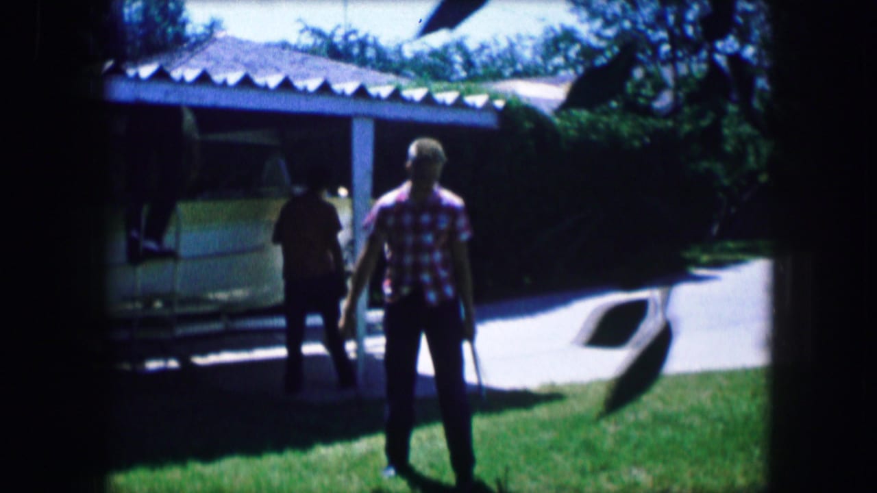 Two people stand on a lawn near a boat stored beside a covered driveway.