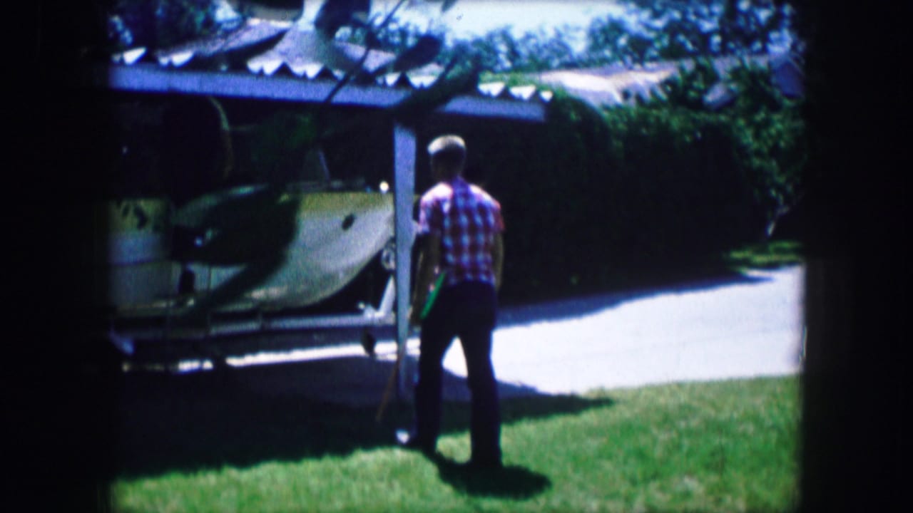 A young man stands on grass beside a boat stored near a driveway.