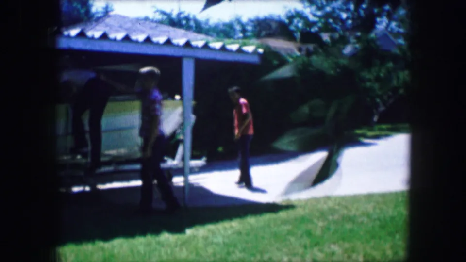 Home-movie frame from Lewisville, Texas showing several friends around a stored boat in a sunny backyard driveway area.