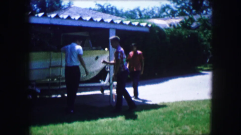 Three people gather near a stored boat in a 1965 Lewisville backyard, with lawn, driveway, and carport visible.