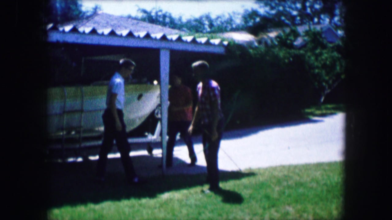 1965 Lewisville, Texas home-movie frame showing three young men near a carport or house as one prepares a bow on a suburban lawn.