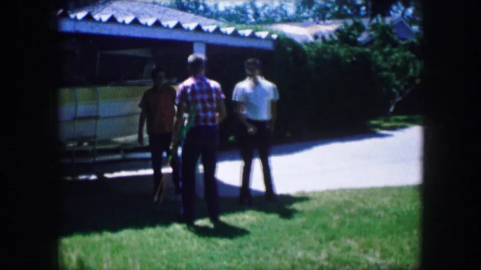 1965 Lewisville, Texas frame with three young men in casual shirts standing on a lawn near a shaded house or carport during archery practice.