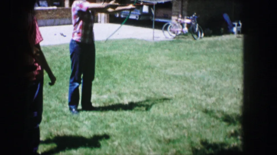 1965 Lewisville, Texas home-movie frame showing a young man with a bow on a suburban lawn while bicycles and a driveway sit in the background.