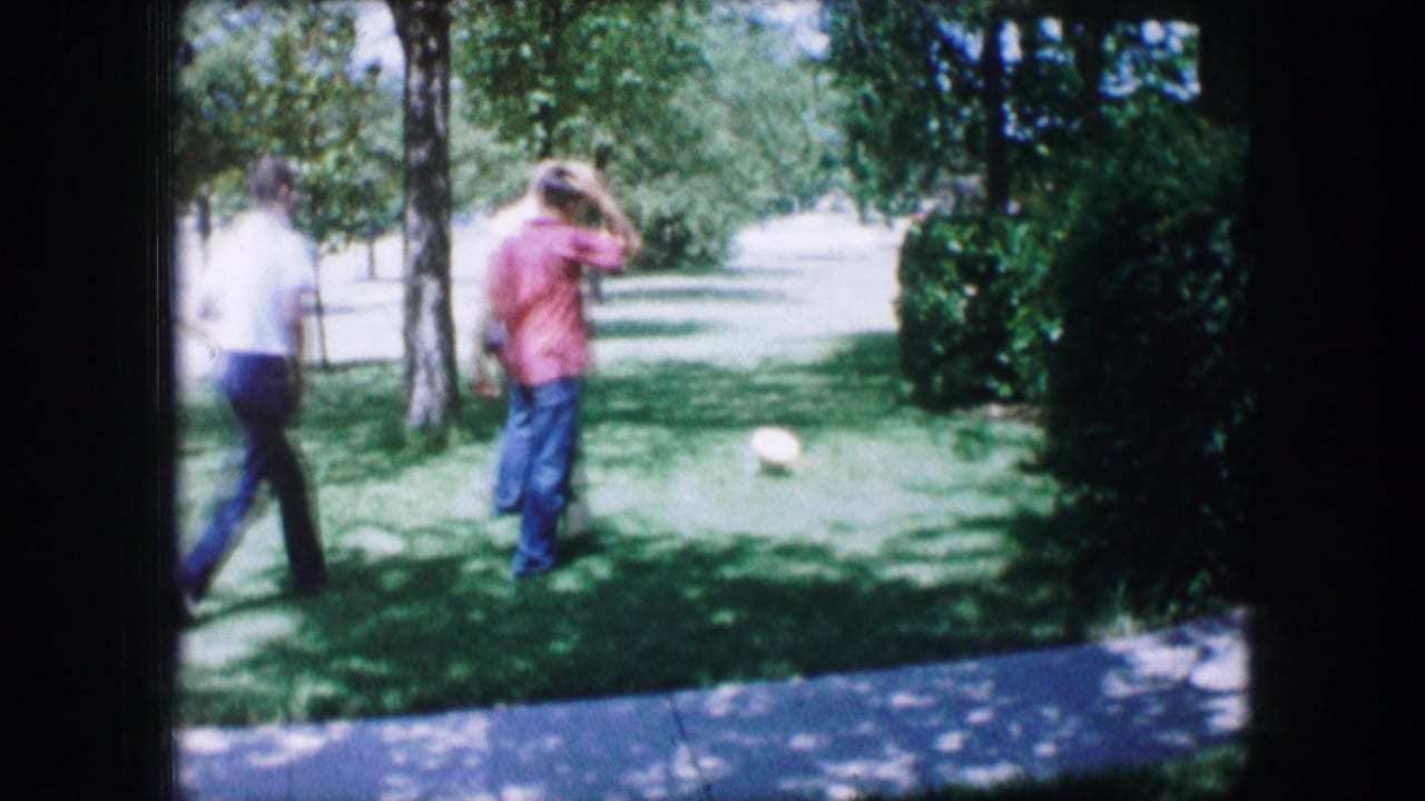 In 1965 Lewisville, Texas home-movie footage, two adult men cross a grassy apartment-area lawn toward a white ball under trees.