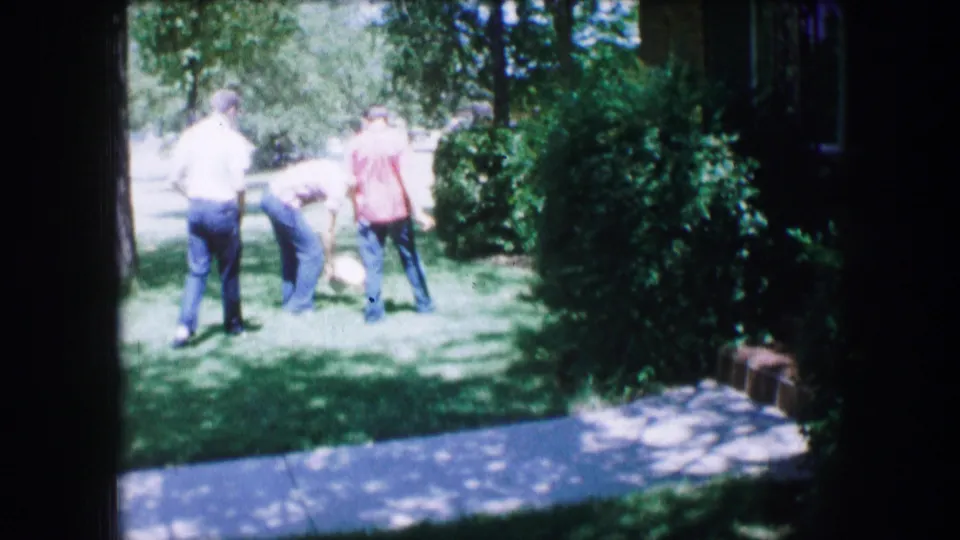 Three adult men stand and bend near a white ball on a grassy lawn by shrubs and a sidewalk in 1965 Lewisville, Texas footage.