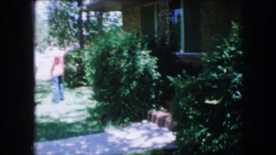 A man in a red shirt stands near dense shrubs, lawn, sidewalk, and a brick apartment exterior in the supplied 1965 Lewisville footage.