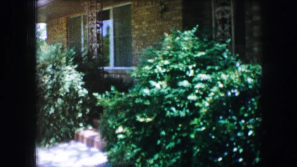 The camera frames shrubs, sidewalk, and a brick apartment facade with windows in 1965 Lewisville, Texas home-movie footage.