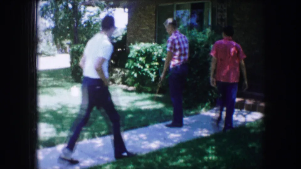 Three casually dressed figures move along a front walkway outside a brick house in this 1965 Lewisville, Texas home-movie frame.