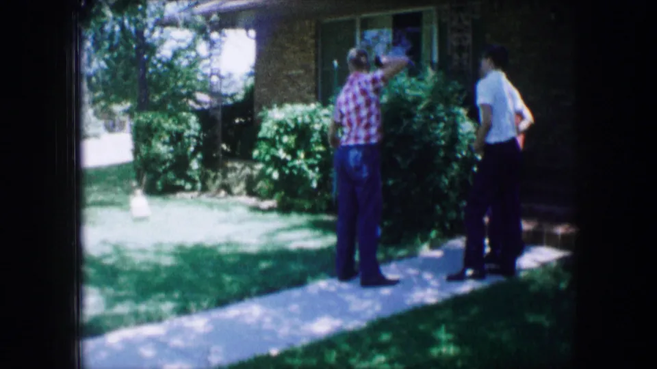Three figures stand near the shrubs and front path of a brick suburban house in 1965 Lewisville, Texas footage.