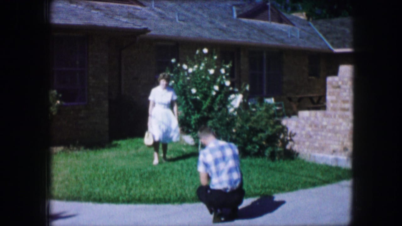 In Lewisville, Texas, 1965, a woman in a white dress approaches a crouching man on a sunny lawn outside a brick home.