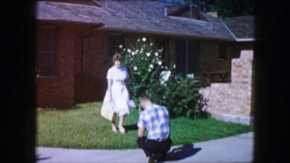 The woman in a white dress moves closer to the crouching man, with a brick house, lawn, and white flowers behind them.