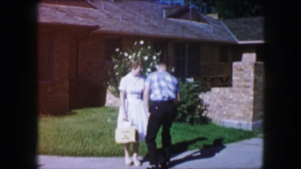 The man rises or steps in beside the woman on the lawn, with the 1965 Lewisville brick home exterior in the background.