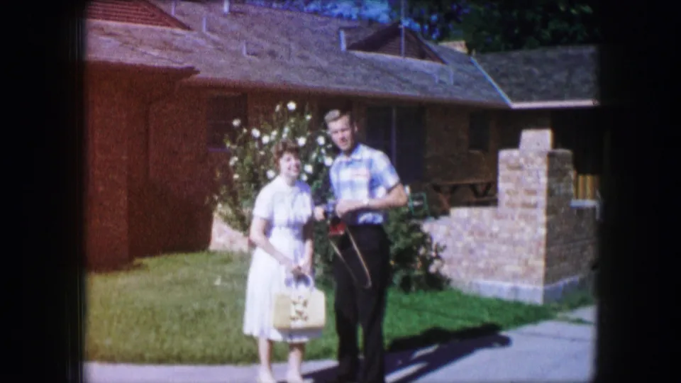 A woman in a white dress and a man in a plaid shirt stand together on a Lewisville lawn, facing the camera in 1965 home-movie footage.