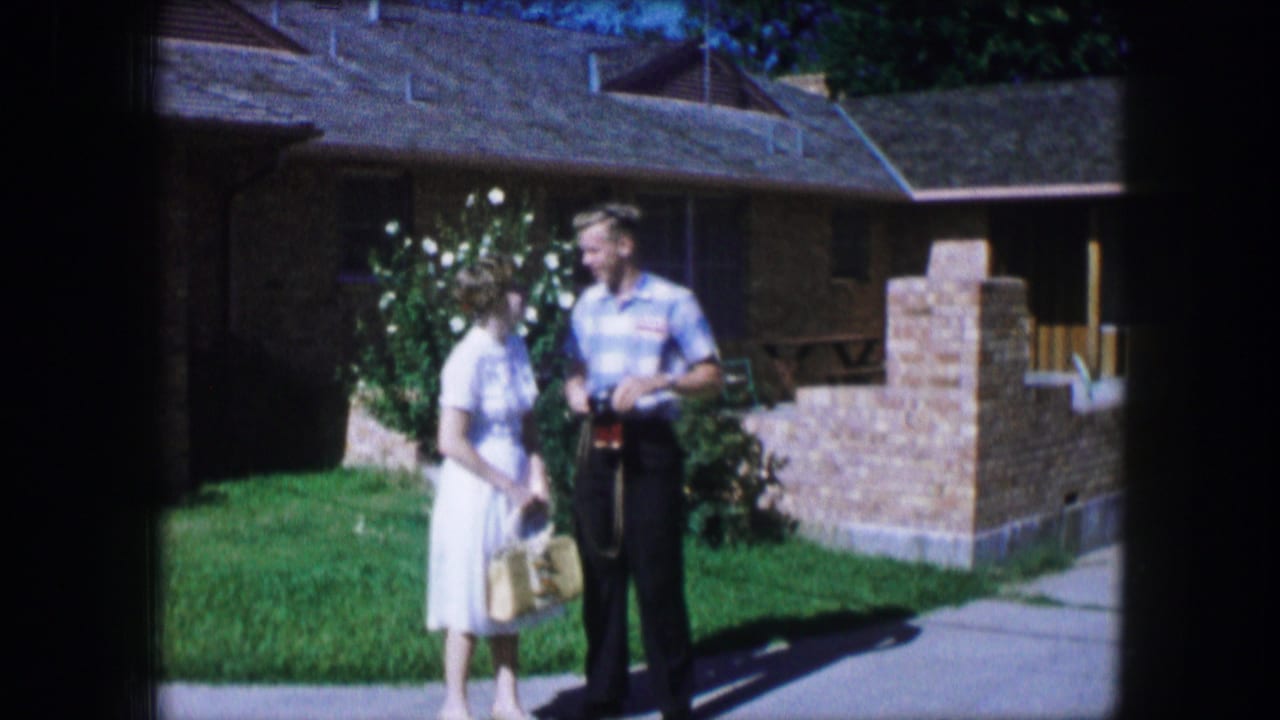 Couple standing on a lawn near a brick house, with the man holding a camera.