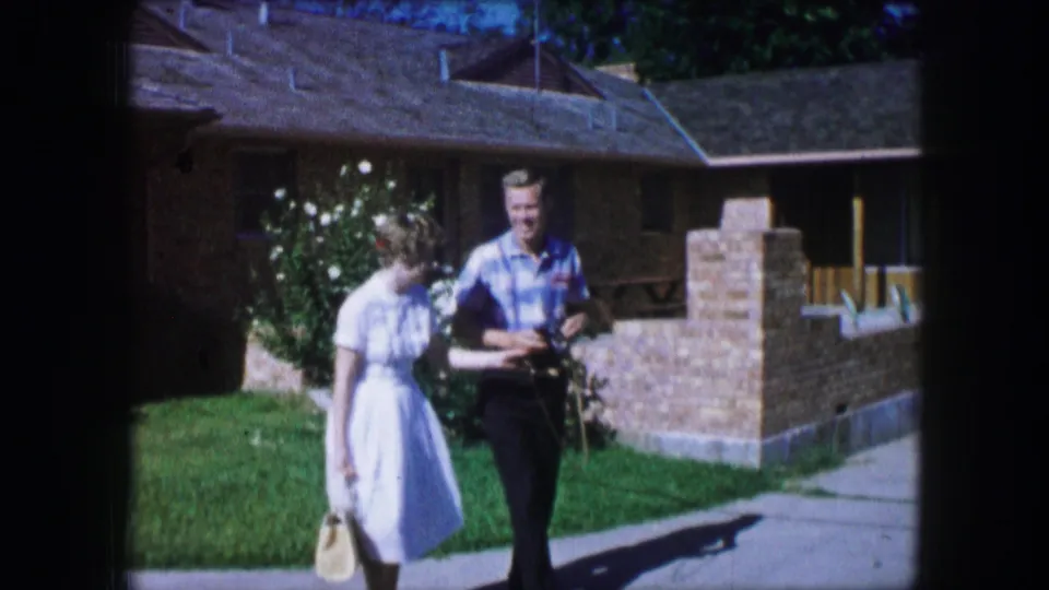 The couple walks across a sunny residential lawn in Lewisville, Texas, with a camera and handbag visible.