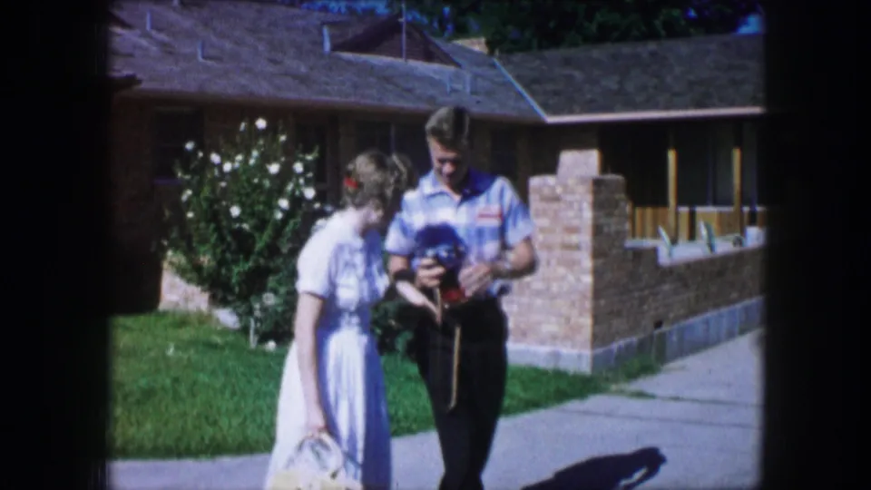 A 1965 Lewisville home-movie frame shows the couple close together while the man checks a small camera.