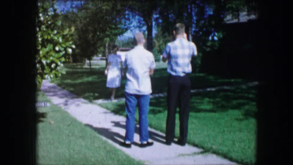 Two adults are seen from behind on a Lewisville residential sidewalk while a woman walks ahead through the yard.