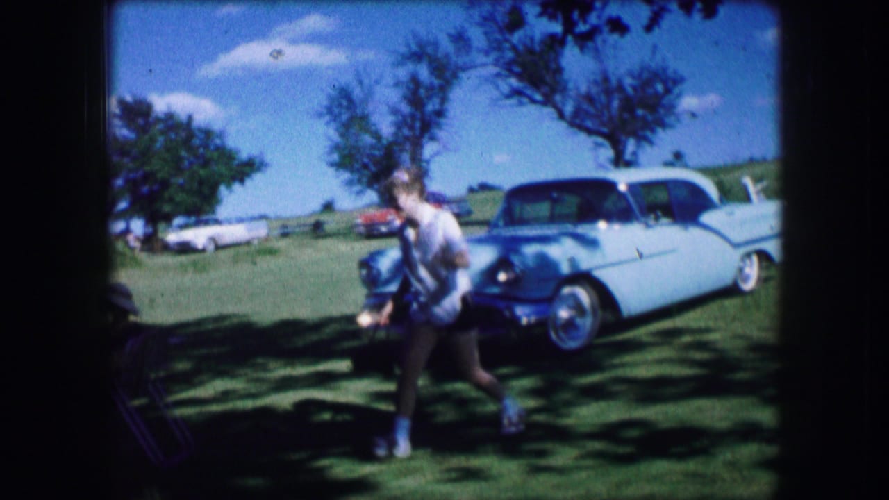In Lewisville, Texas, 1965 home-movie footage, a woman walks through a grassy shaded area near folding chairs and a white classic car.