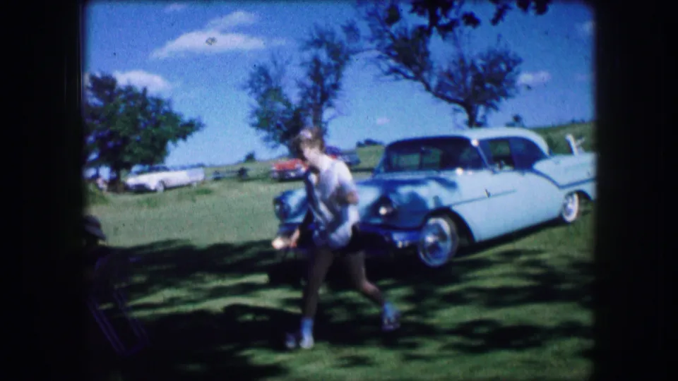 In Lewisville, Texas, 1965 home-movie footage, a woman walks through a grassy shaded area near folding chairs and a white classic car.