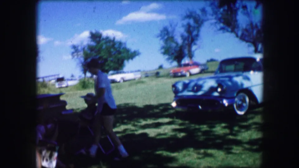 The woman moves through shade beside lawn chairs while a white mid-century car sits on the grass in the background.
