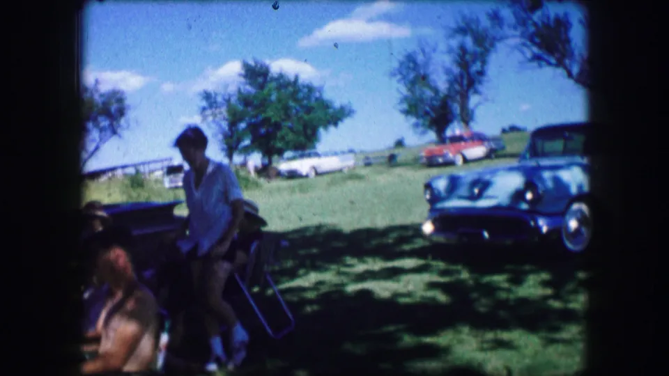 In the shaded foreground, the woman nears seated adults while the white classic car and other parked cars remain visible on the sunny field.