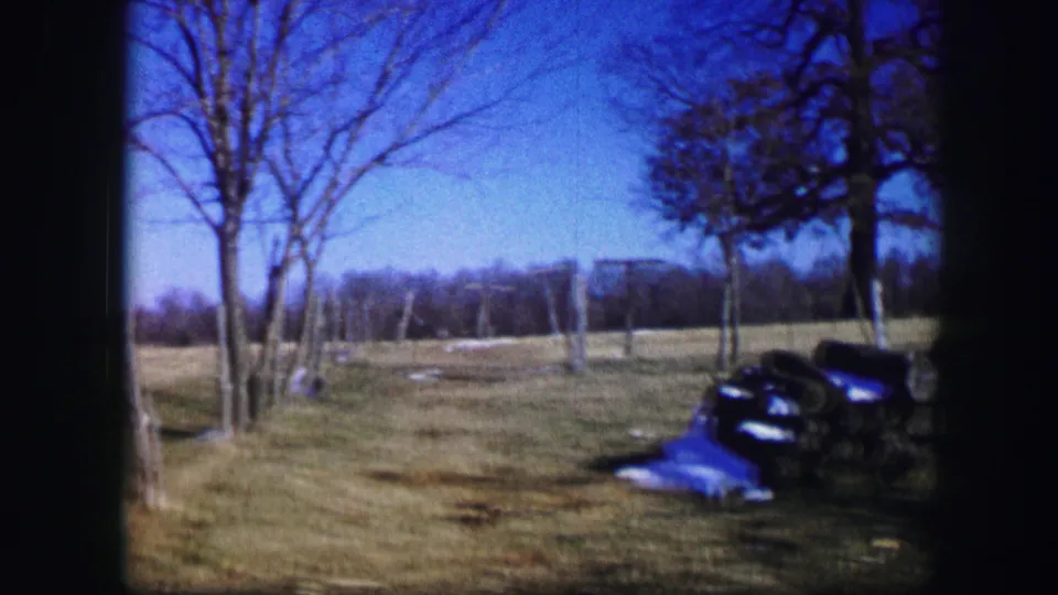 1967 Kirksville 8mm view across a rural yard with bare trees, fence posts, open field, dark equipment, and a blue tarp.